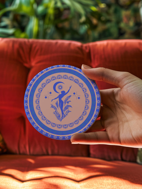 Round ceramic coaster featuring a silhouetted dancing figure reaching toward a crescent moon, surrounded by concentric rings of stars and floral patterns in blue on peach background